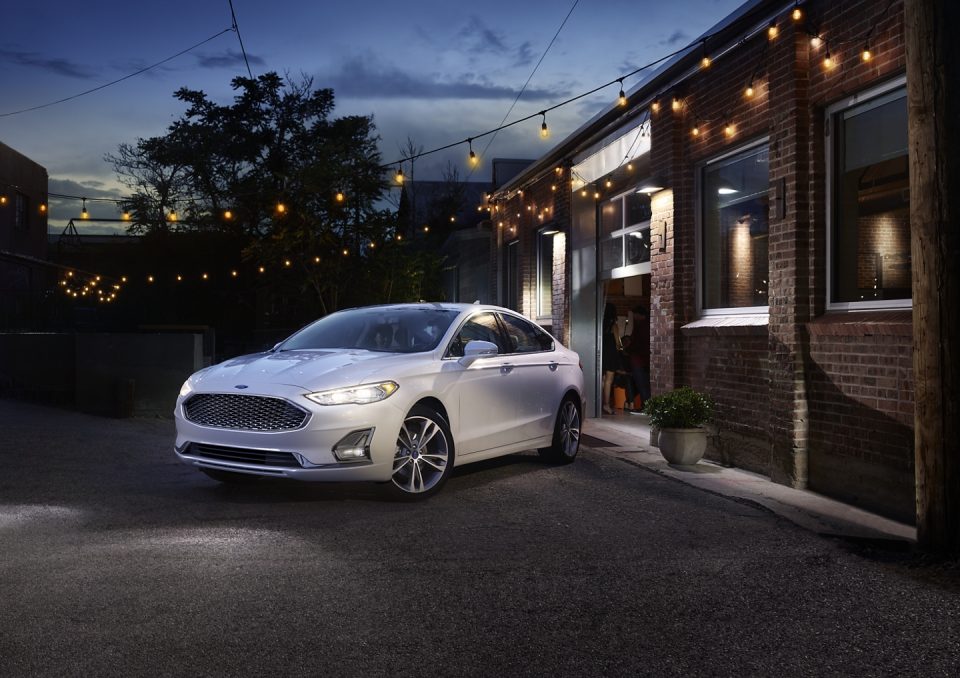 A silver 2020 Ford Fusion against a brick building at night, string lights overhead of the car illuminate the scene.