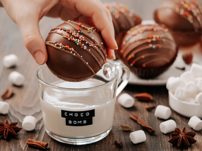 Chocolate cocoa bomb in hand near glass cup with plant-based milk and choco bomb text. Ball made from milk chocolate with marshmallow. Stylish orange toned image of trendy winter hot chocolate drink